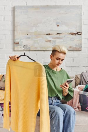 joven mujer tatuada sosteniendo jersey amarillo y mirando el teléfono móvil mientras está sentado en el sofá cerca de artículos de armario, clasificación de ropa, intercambio en línea, vida sostenible y concepto de consumismo consciente