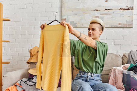 joven mujer tatuada sonriendo y mirando el jersey amarillo de moda mientras está sentado en el sofá cerca de la tienda de segunda mano encuentra en la sala de estar moderna en casa, vida sostenible y concepto de consumismo consciente