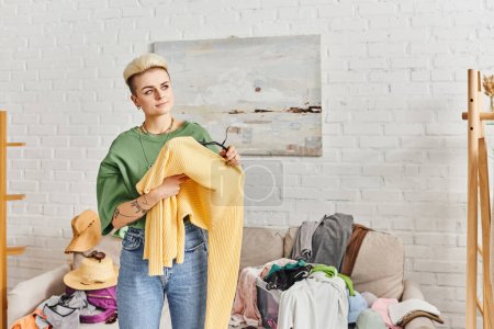 conscious decluttering, clothing sorting, smiling tattooed woman holding yellow jumper and looking away near couch with wardrobe items at home, sustainable living and mindful consumerism concept