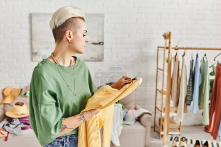 stylish tattooed woman looking at yellow jumper near rack with clothing and footwear in modern living room, wardrobe items sorting, sustainable living and mindful consumerism concept