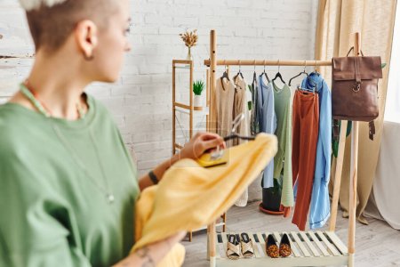 verschwommene Frau mit gelbem Pullover blickt auf Regal mit Kleiderbügeln, Kleidung, Ledertasche und Schuhen im modernen Wohnzimmer, Kleidersortierung, nachhaltiges Leben und achtsames Konsumkonzept