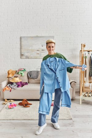 wardrobe and thrift store finds sorting, joyful tattooed woman showing blue pajamas and standing in modern living near couch and rack with clothes, sustainable living and mindful consumerism concept