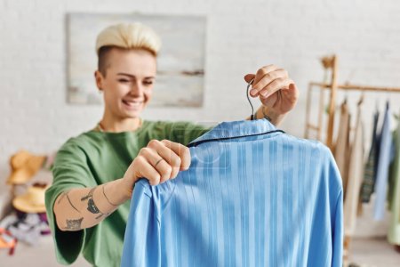 Photo for Thrift clothes, clothing sorting, pleased tattooed woman holding hanger with blue pajamas in living room at home on blurred background, sustainable living and mindful consumerism concept - Royalty Free Image