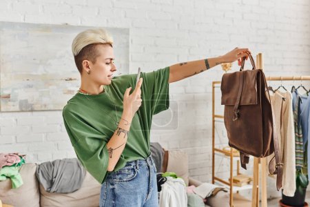 sorting clothes, online exchange, tattooed woman taking photo of leather bag near couch and rack with clothes in modern living room, sustainable living and mindful consumerism concept