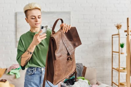 swap on virtual marketplace, young woman with tattoo and trendy hairstyle taking photo of leather bag on mobile phone near couch with clothing, sustainable living and mindful consumerism concept