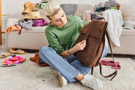 clothing decluttering, tattooed woman with trendy hairstyle sitting on floor with leather bag near wardrobe garments on couch in living room, sustainable living and mindful consumerism concept