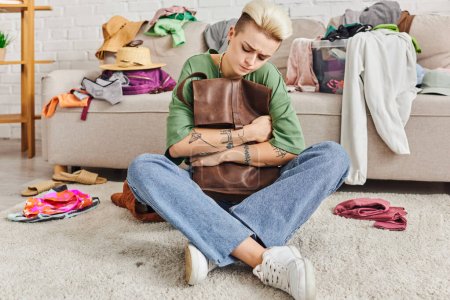 Photo for Sorting pre-loved items, home decluttering, thoughtful tattooed woman sitting on floor with leather bag near couch with clothes at home, sustainable living and mindful consumerism concept - Royalty Free Image