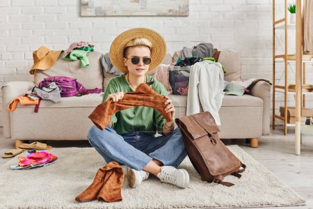 vêtements d'épargne, femme à la mode en chapeau de paille et lunettes de soleil assis sur le sol près du sac en cuir et tenant des bottes en daim près du canapé dans le salon, la mode durable et le concept de consumérisme conscient