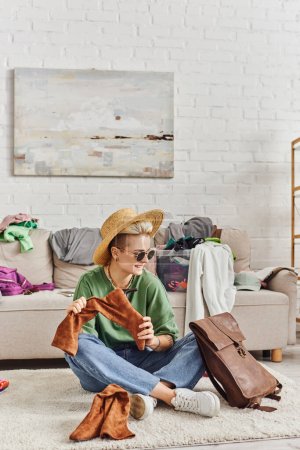 smiling and fashionable woman in straw hat and sunglasses sitting on floor with leather bag and suede boots near couch in living room, sustainable fashion and mindful consumerism concept