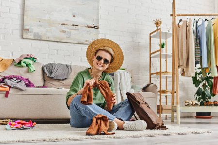 Téléchargez les photos : Tri des vêtements, femme tatouée ravie en chapeau de paille et lunettes de soleil regardant la caméra près de bottes en daim et sac en cuir sur le sol à la maison, la mode durable et le concept de consumérisme conscient - en image libre de droit