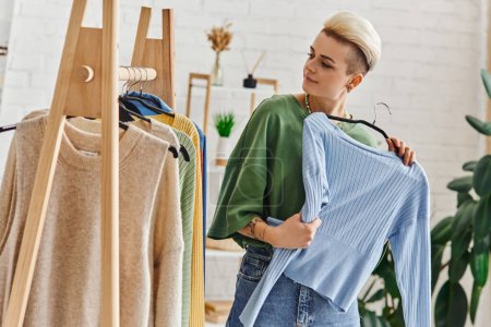 decluttering conscient, femme tatouée heureux tenant cardigan bleu et regardant rack avec des vêtements à la mode sur cintres dans le salon, mode durable et concept de consumérisme conscient