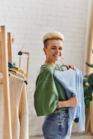excited tattooed woman with trendy hairstyle holding blue cardigan and smiling at camera near rack with thrift store finds on hangers at home, sustainable fashion and mindful consumerism concept