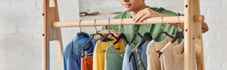 partial view of smiling and tattooed woman standing near rack with fashionable casual garments on hangers at home, sustainable fashion and mindful consumerism concept, banner