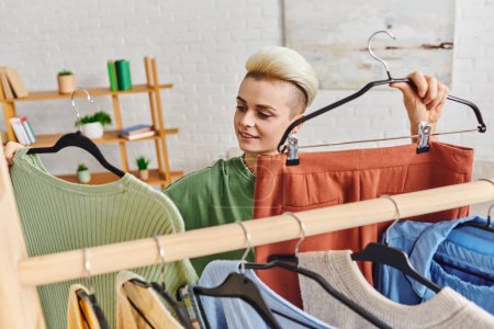 positive young woman holding hangers with pants and jumper near rack with wardrobe garments, clothing sorting, conscious decluttering, sustainable fashion and mindful consumerism concept
