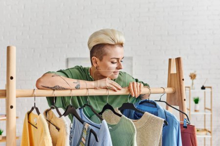 positive and tattooed woman with trendy hairstyle leaning on rack and looking at casual clothing on hangers at home, pre-loved items, sustainable fashion and mindful consumerism concept