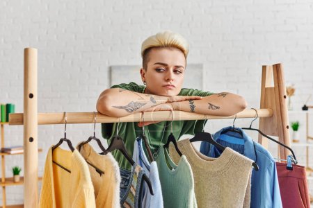 thoughtful and sad tattooed woman learning on rack with fashionable casual clothes on hangers in modern living room at home, fashion and mindful consumerism concept