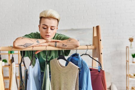 Photo for Wardrobe items sorting, dreamy tattooed woman with trendy hairstyle leaning on rack with fashionable casual clothes on hangers, sustainable fashion and mindful consumerism concept - Royalty Free Image