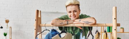 Photo for Young tattooed woman with trendy hairstyle and happy face looking at camera and leaning on rack with hangers and stylish clothes at home, sustainable fashion and mindful consumerism concept, banner - Royalty Free Image