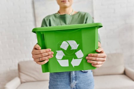 enfoque en la caja de plástico verde con el cartel de reciclaje en las manos de la mujer recortada de pie en casa sobre un fondo borroso, la vida sostenible y el concepto de hábitos ecológicos