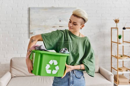responsabilidad social, mujer joven y tatuada sosteniendo caja de reciclaje verde con prendas de vestir en la sala de estar, emoción positiva, vida sostenible y concepto de hábitos ecológicos