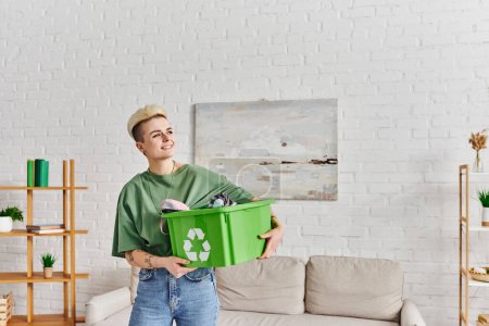 fröhliche tätowierte Frau mit Kleidung in Plastikbox mit Recyclingschild in modernem Wohnzimmer mit grünen Pflanzen auf Regalen, nachhaltigem Leben und umweltfreundlichem Gewohnheitskonzept