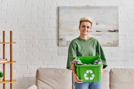 zufriedene, tätowierte junge Frau in Freizeitkleidung, grüne Recyclingbox mit Kleidungsstücken in der Hand und Blick in die Kamera im modernen Wohnzimmer, nachhaltiges Leben und umweltfreundliches Gewohnheitskonzept