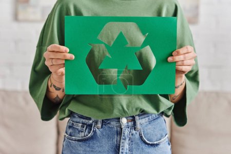 Green Living, Teilbild einer jungen tätowierten Frau in Freizeitkleidung mit grüner Papierkarte mit ausgeschnittenem Recyclingschild zu Hause, nachhaltiges und umweltbewusstes Lifestylekonzept