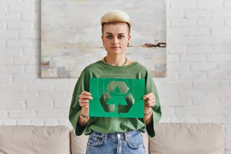 young woman in stylish casual clothes, with trendy hairstyle holding green paper card with cutout recycling symbol and looking at camera, sustainable and environmentally conscious lifestyle concept