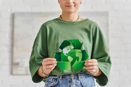 Téléchargez les photos : Mode de vie éco-conscient, vue partielle de la femme tatouée souriante dans des vêtements décontractés tenant le symbole de recyclage vert partout dans le monde à la maison, concept de vie durable et de sensibilisation à l'environnement - en image libre de droit