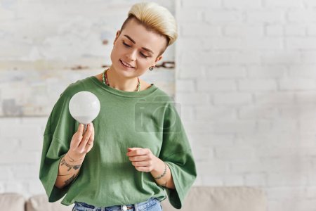 cheerful young woman in casual clothes looking at energy saving light bulb at home, trendy hairstyle, tattoo, positive emotion, sustainable lifestyle and environmentally conscious concept