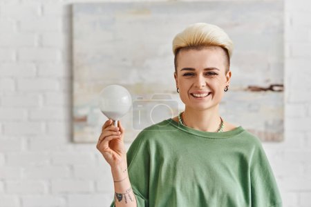 ecological efficiency, joyful tattooed woman with trendy hairstyle holding energy saving light bulb and looking at camera in living room, sustainable lifestyle and environmentally conscious concept