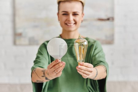 Photo for Cheerful, young and tattooed woman in casual clothes showing energy saving light bulbs and looking at camera, environmentally conscious, sustainable lifestyle and environmentally conscious concept - Royalty Free Image