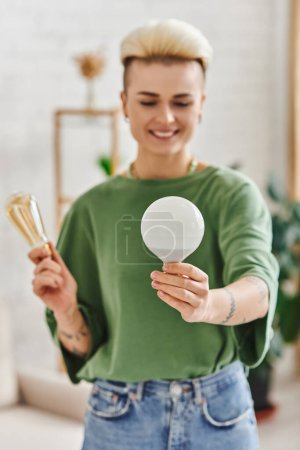 Photo for Young and tattooed woman in casual clothes holding energy saving light bulbs and smiling in living room on blurred background,  sustainable lifestyle and environmentally conscious concept - Royalty Free Image