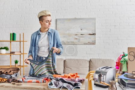 sharing economy, tattooed and stylish woman with knitted tank-top near table with second-hand clothing and electric toaster, belongings exchange, sustainable living and mindful consumerism concept