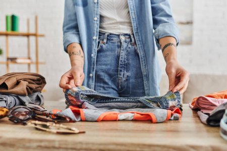 Teilbild einer jungen tätowierten Frau in Freizeitkleidung, die Kleiderschränke auf dem Wohnzimmertisch sortiert, Tauschbörse, umweltfreundliche Tauschgeschäfte, nachhaltiges Leben und bewusstes Konsumkonzept