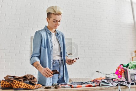 smiling and stylish tattooed woman holding sunglasses near table with garments while sorting clothing and pre-loved items for exchange market, sustainable living and mindful consumerism concept