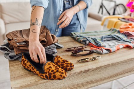 cropped view of tattooed woman placing animal print shoes on table with second-hand items and sunglasses, ethical consumption, exchange, sustainable living and mindful consumerism concept