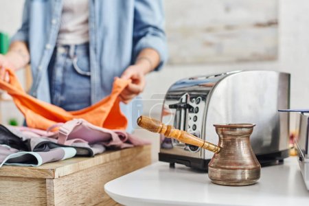 partial view of young woman sorting clothes near electric toaster and cezve, eco-friendly swaps, exchange market, blurred background, sustainable living and promoting circular economy concept