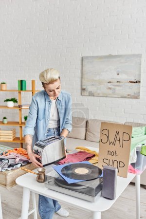 Photo for Casually styled and tattooed woman holding electric toaster near vinyl record player, cezve, pre-loved items and swap not shop card in living room, sustainable living and circular economy concept - Royalty Free Image