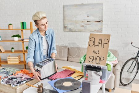 young tattooed woman with electric toaster near vinyl record player, second-hand clothes and carton card with swap not shop lettering at home, sustainable living and circular economy concept