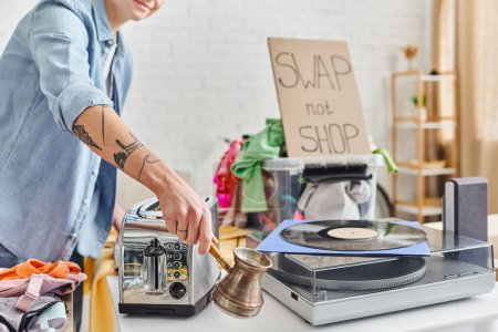partial view of tattooed woman holding cezve near electric toaster, vinyl record player, clothes and card with swap not shop lettering at home, sustainable living and circular economy concept