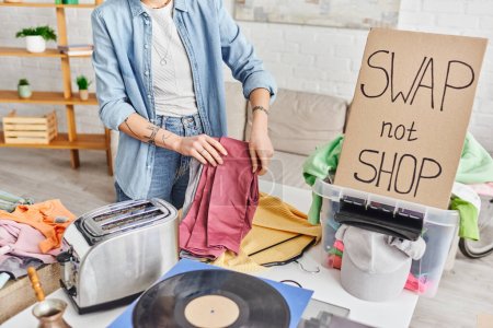 cropped view of tattooed woman with pants near vinyl disc, electric toaster, wardrobe items, plastic box and swap not shop card, exchange market, sustainable living and circular economy concept