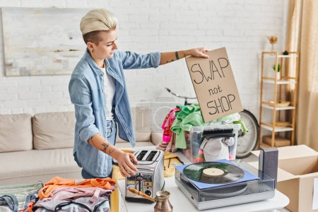 Photo for Tattooed woman with trendy hairstyle holding swap not shop card near vinyl record player, electric toaster, cezve and wardrobe clothes in living room, sustainable living and circular economy concept - Royalty Free Image