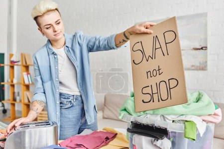 tattooed woman in casual clothes holding swap not shop card near electric toaster, plastic container and clothes in modern living room, sustainable living and circular economy concept