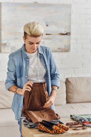 resale market, young tattooed woman in stylish casual clothes folding leather pants near animal print shoes and garments on table at home, sustainable living and mindful consumerism concept