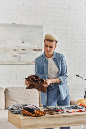 young and tattooed woman in casual clothes, with trendy hairstyle sorting wardrobe items on table in modern living room, clothing swap, sustainable living and conscious lifestyle concept