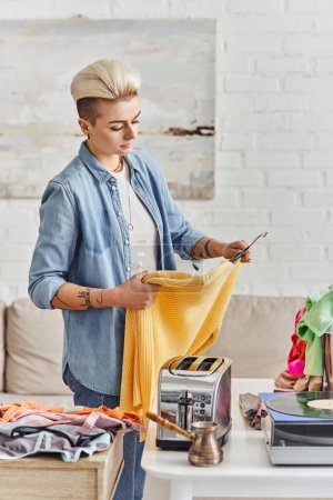 Photo for Exchange market, tattooed woman with trendy hairstyle holding yellow jumper near table with electric toaster, vinyl record player and clothes, sustainable living and mindful consumerism concept - Royalty Free Image