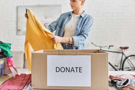 mujer tatuada en ropa casual mirando jersey amarillo cerca de prendas de armario y caja de cartón con letras de donar, fondo borroso, vida sostenible y concepto de responsabilidad social