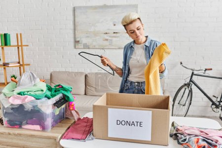 tattooed woman with trendy hairstyle holding hanger and yellow jumper near plastic container with clothing and donation box in living room, sustainable living and social responsibility concept