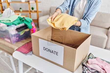 vista parcial de una mujer joven y tatuada sosteniendo un jersey amarillo encima de una caja de cartón con letras de donaciones cerca de un contenedor de plástico con ropa, vida sostenible y concepto de responsabilidad social
